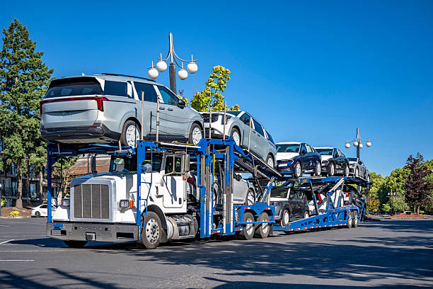 A large car carrier truck, part of a Car Transport Naples, FL service, transports multiple new SUVs and sedans on a sunny day, driving down a wide street lined with trees and vintage street lamps.